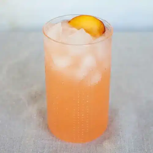 A tall glass and pitcher of vibrant peach lemonade, garnished with a fresh peach slice and a sprig of mint.