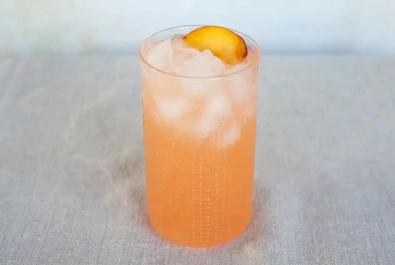 A tall glass and pitcher of vibrant peach lemonade, garnished with a fresh peach slice and a sprig of mint.