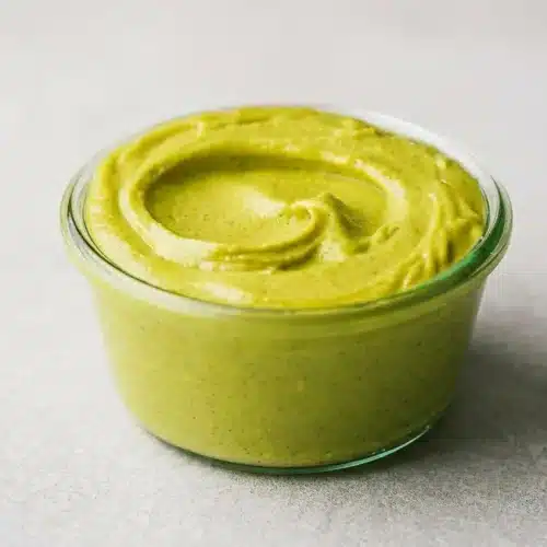 A small glass jar of vibrant green Pistachio Cream with a smooth, velvety texture, a small spoon resting on top, and pistachios scattered on