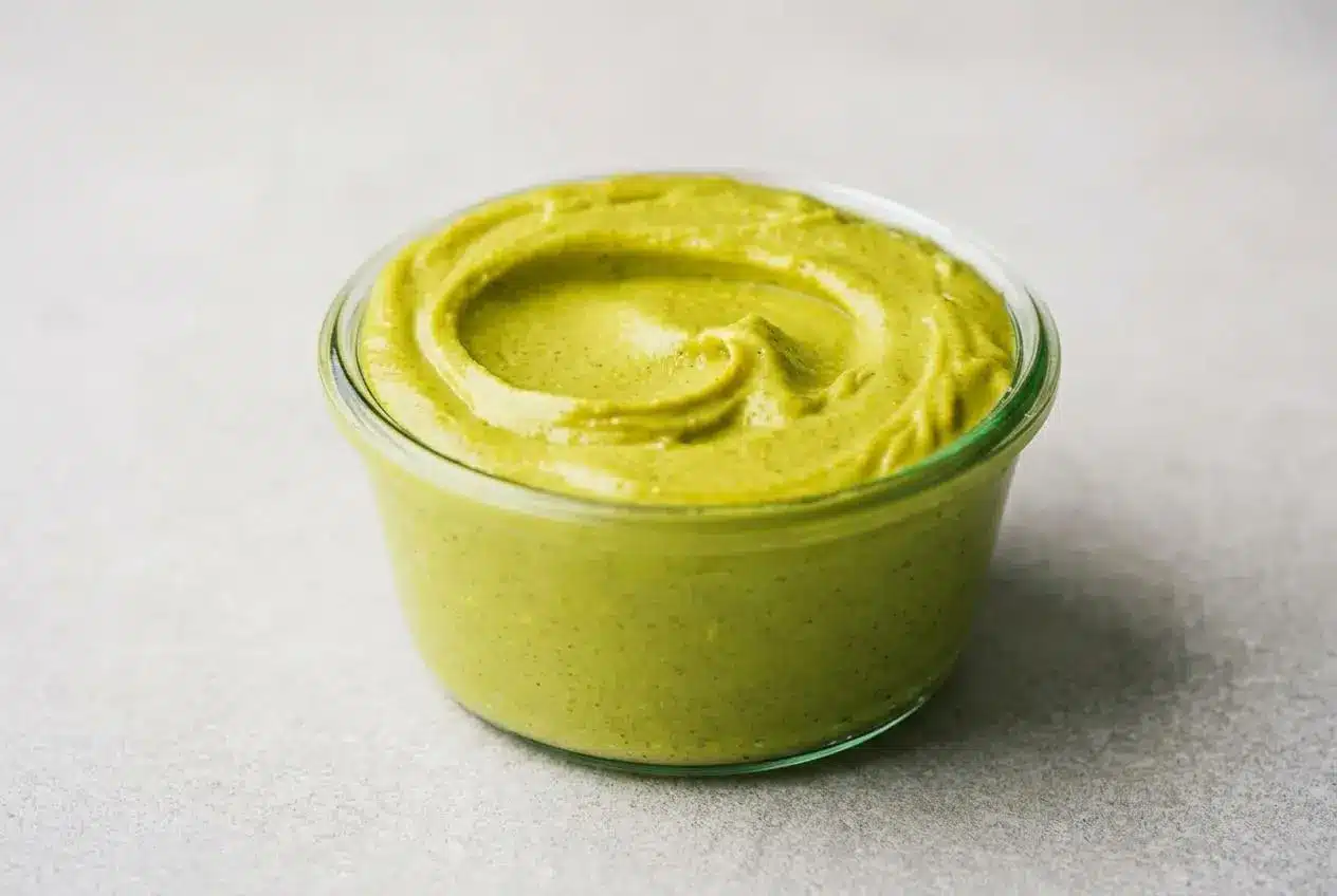 A small glass jar of vibrant green Pistachio Cream with a smooth, velvety texture, a small spoon resting on top, and pistachios scattered on