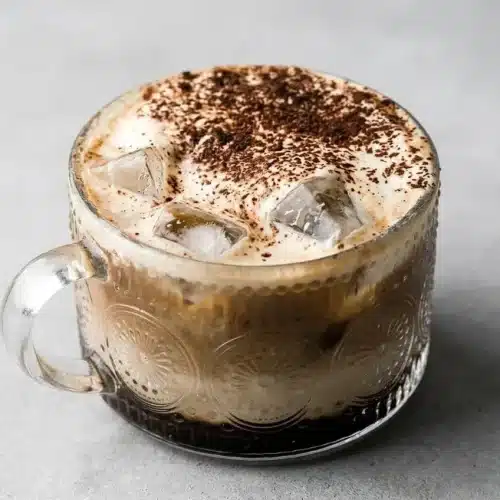 Iced Tiramisu Latte in a tall glass showing distinct layers and a creamy mascarpone foam topping dusted with cocoa.