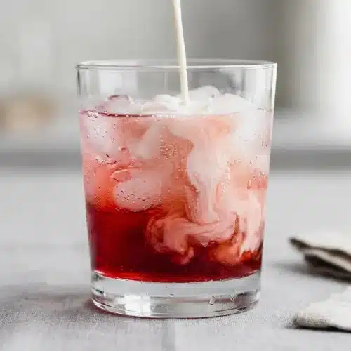 A tall glass of raspberry Italian Cream Soda with ice, topped with whipped cream and a cherry, showing creamy swirls.