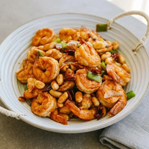 A white bowl filled with kung pao shrimp, showing tender shrimp, crunchy peanuts, and scallions coated in a glossy, savory sauce.