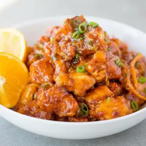 A white bowl of crispy orange peel chicken with a glossy, tangy sauce, garnished with fresh green onion and julienned orange peel.