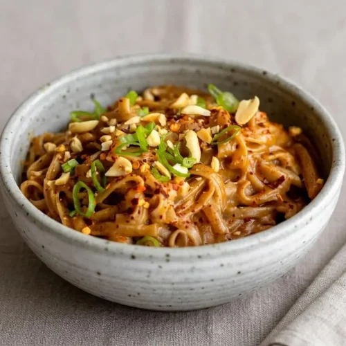 A bowl of Peanut Butter Noodles with a creamy sauce, lifted by chopsticks and topped with crushed peanuts and scallions.