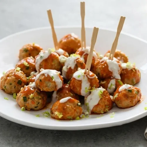 A white bowl of buffalo chicken meatballs coated in a glossy orange sauce, garnished with scallions and served with blue cheese dip.