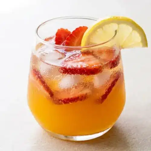 A tall glass of vibrant orange strawberry mango lemonade filled with ice, a lemon wheel, and a fresh strawberry slice.