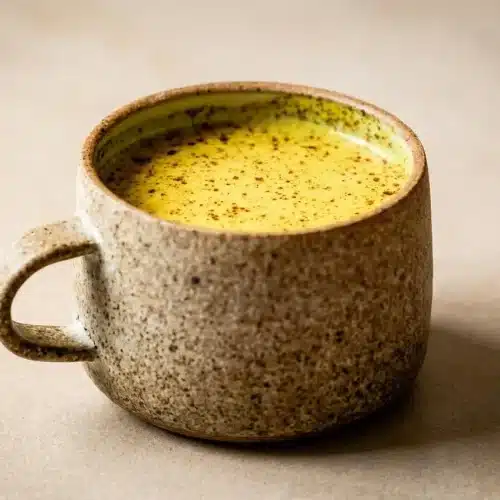 A steaming mug of creamy Turmeric Golden Milk Recipe, garnished with a light dusting of cinnamon on a dark surface.