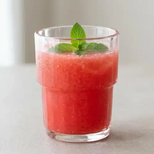 A glass and pitcher of vibrant pink watermelon juice recipe, garnished with fresh mint and a lime wedge for a refreshing summer drink.