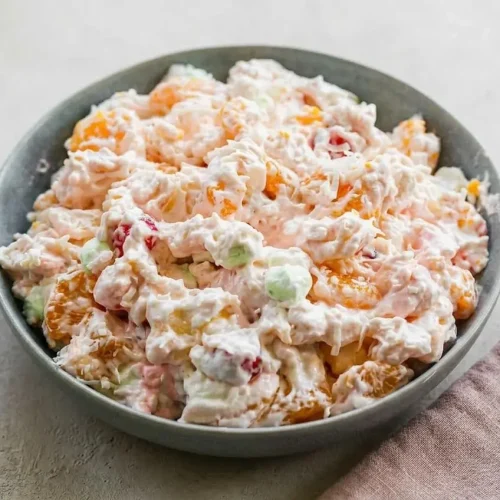 A large glass bowl of creamy ambrosia fruit salad, filled with mandarin oranges, cherries, and colorful mini marshmallows.