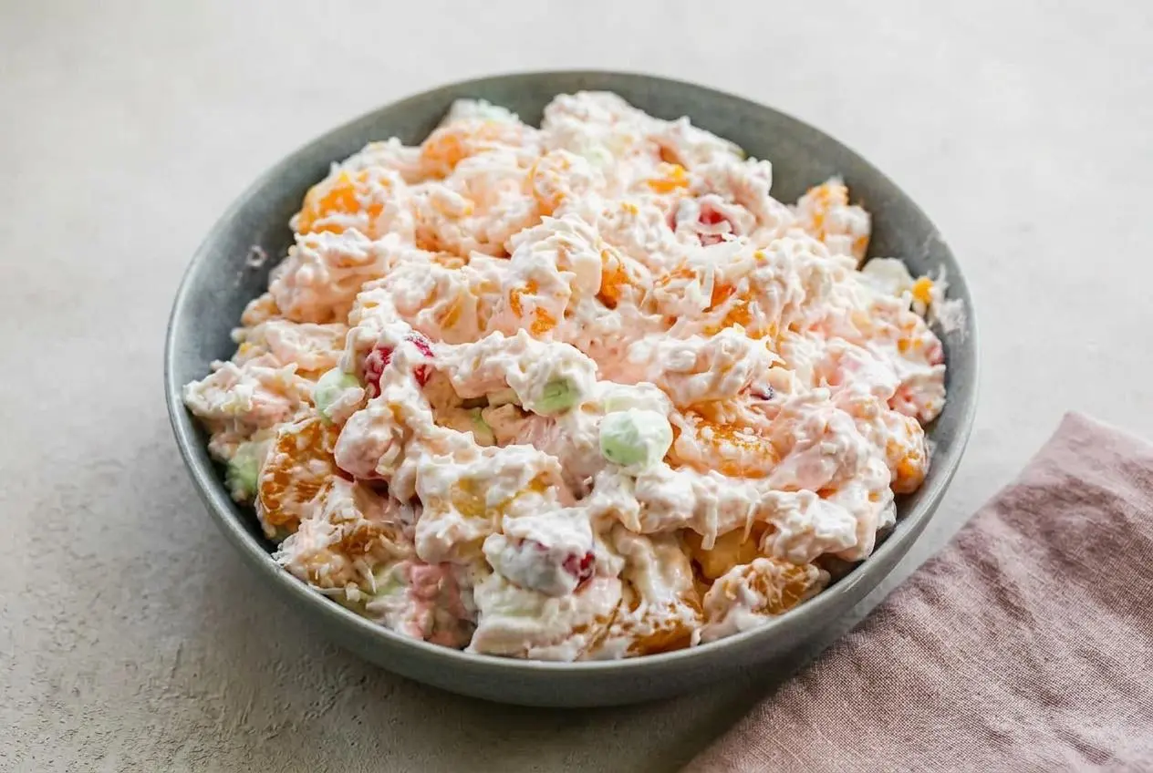 A large glass bowl of creamy ambrosia fruit salad, filled with mandarin oranges, cherries, and colorful mini marshmallows.