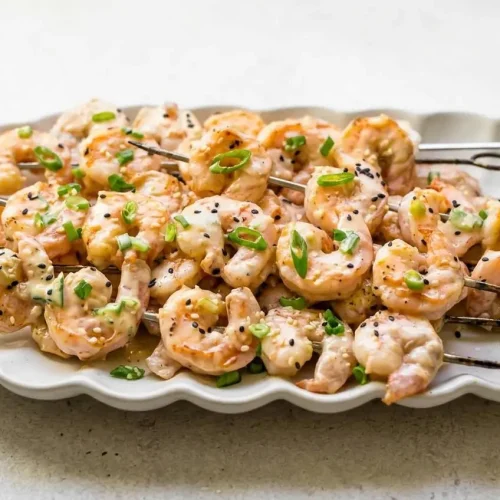 A white platter of Bang Bang Shrimp Skewers, grilled with char marks, coated in a creamy sauce, and sprinkled with scallions.