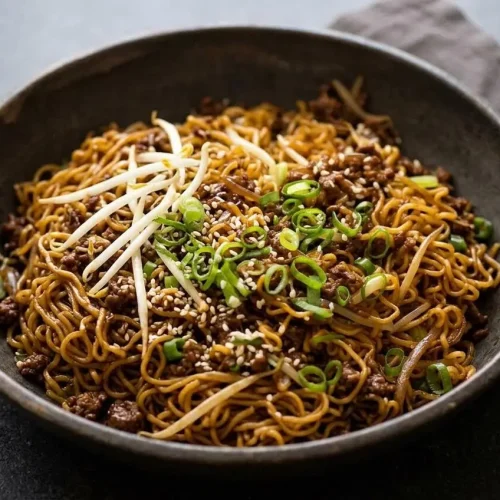 A bowl of beef ramen noodles with caramelized ground beef and a glossy savory-sweet sauce, garnished with green onions and sesame seeds.