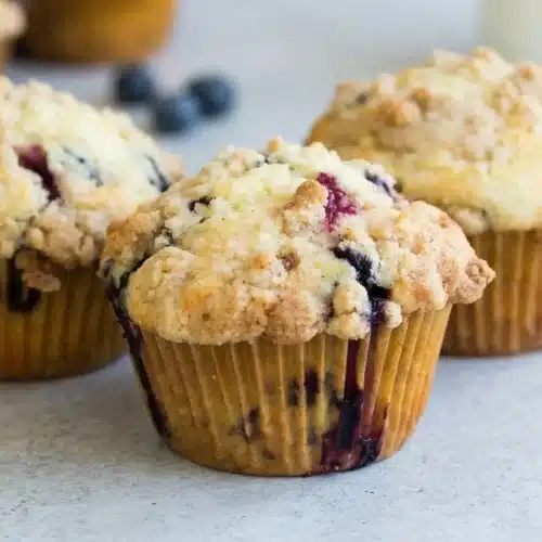 A dozen blueberry muffins from this recipe in a metal muffin tin, showing their tall, golden-brown tops with a crunchy streusel.