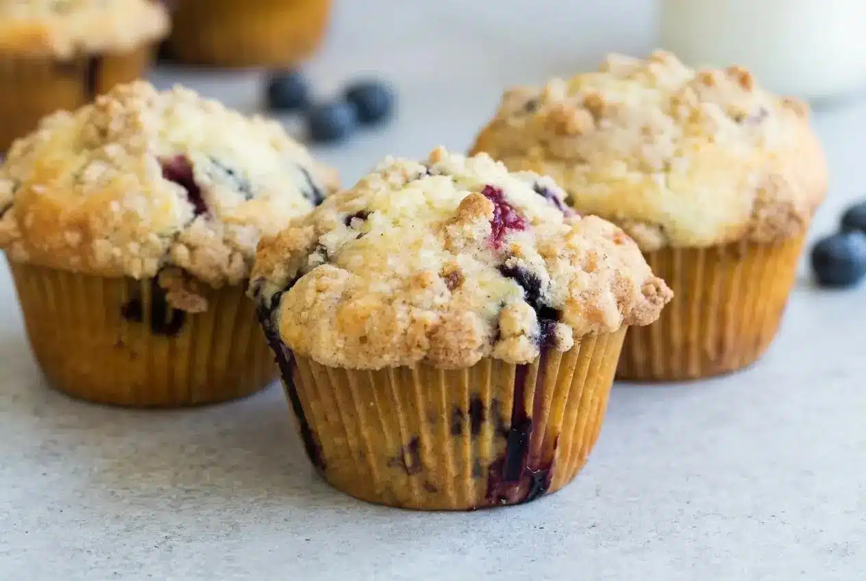 A dozen blueberry muffins from this recipe in a metal muffin tin, showing their tall, golden-brown tops with a crunchy streusel.