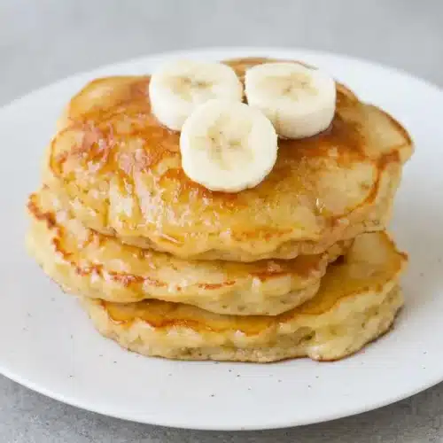 A tall stack of brown sugar banana pancakes on a white plate, topped with a pat of melting butter and a drizzle of maple syrup.