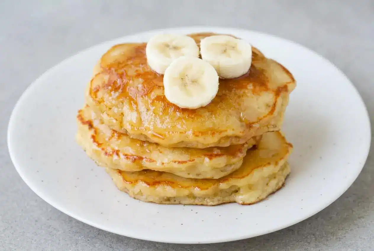 A tall stack of brown sugar banana pancakes on a white plate, topped with a pat of melting butter and a drizzle of maple syrup.