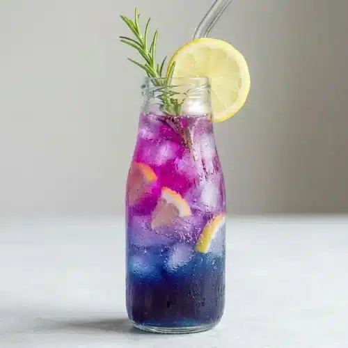 A tall, clear glass of vibrant purple butterfly pea flower lemonade with ice, garnished with a fresh lemon slice and a rosemary sprig.