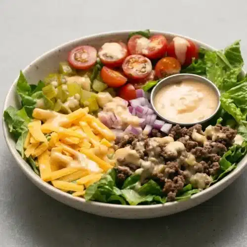 A fresh cheeseburger salad in a white bowl, with seasoned ground beef, shredded cheddar, tomatoes, and pickles over romaine, drizzled with