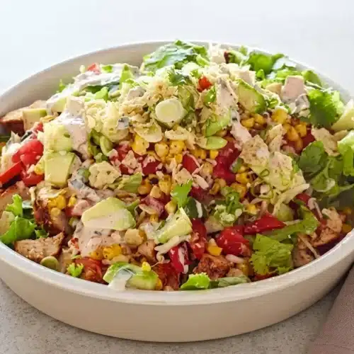 A fresh and hearty chicken taco salad in a white bowl, topped with seasoned chicken, charred corn, tomatoes, avocado, and crushed tortilla