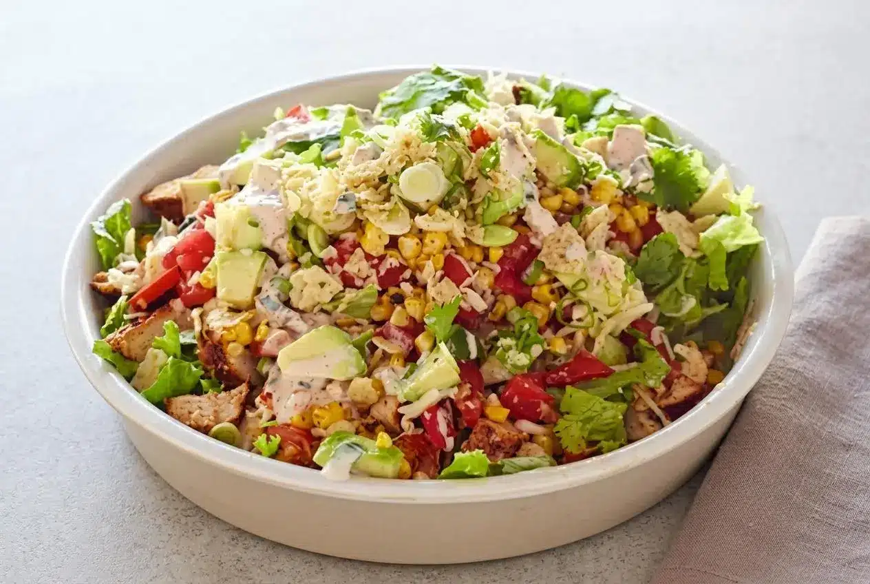 A fresh and hearty chicken taco salad in a white bowl, topped with seasoned chicken, charred corn, tomatoes, avocado, and crushed tortilla