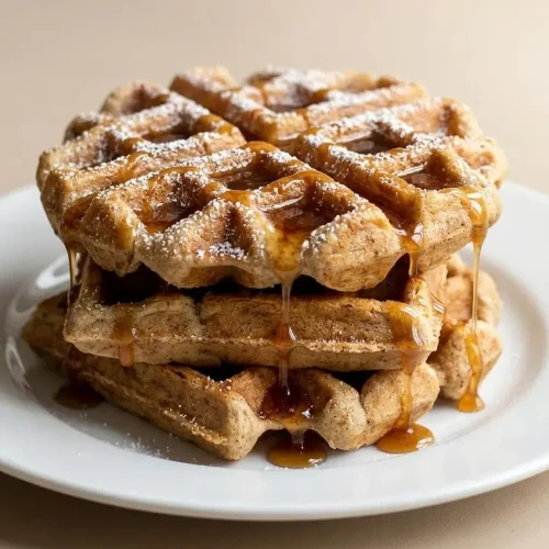 A stack of golden Cinnamon Brown Sugar Waffles on a white plate, with a pat of melting butter on top and maple syrup being poured over.