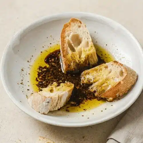 Olive Oil Bread Dip in a shallow white bowl, with glossy pools of balsamic vinegar and herbs, served with a piece of crusty bread dipped in.