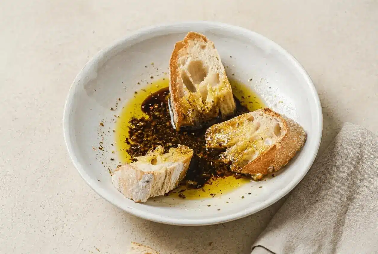 Olive Oil Bread Dip in a shallow white bowl, with glossy pools of balsamic vinegar and herbs, served with a piece of crusty bread dipped in.