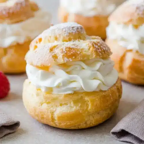 A cream puffs recipe showing several finished puffs on a white plate, one cut open to reveal whipped cream and a raspberry inside, all