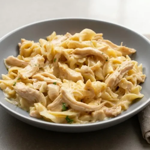 A top-down view of a rich chicken and noodles recipe served in a dark bowl, showing tender shredded chicken and wide egg noodles in a creamy