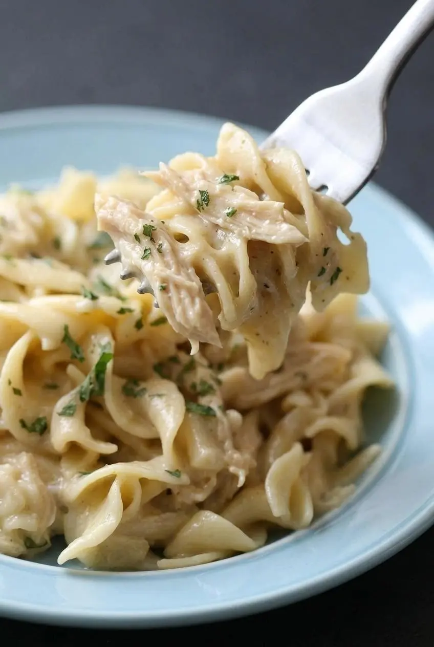 A close-up of a fork lifting creamy chicken and noodles, showing the thick sauce, tender egg noodles, and shredded chicken.