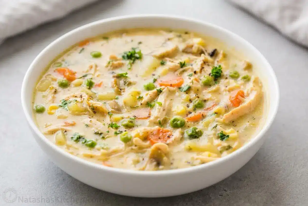 A white bowl of creamy chicken pot pie soup filled with tender chicken, carrots, and peas, garnished with fresh parsley.