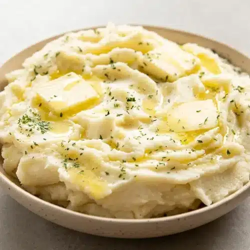A white ceramic bowl filled with creamy Instant Pot mashed potatoes, topped with a melting pat of butter and fresh herbs.