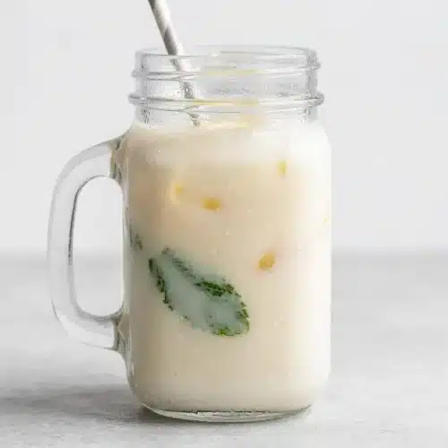A tall glass of creamy lemonade with a velvety smooth texture, garnished with a lemon slice and mint sprig, next to a pitcher.