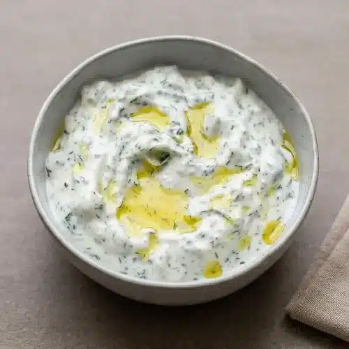A white ceramic bowl of thick and creamy tzatziki sauce recipe, topped with fresh dill and a swirl of golden olive oil.