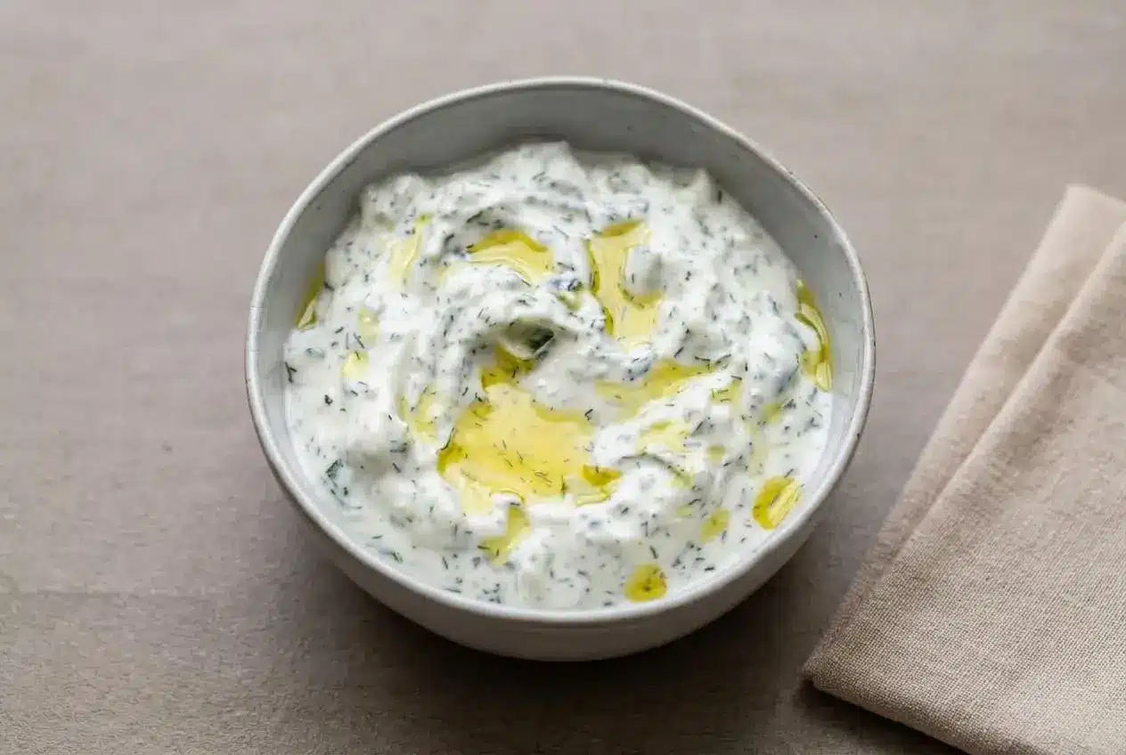 A white ceramic bowl of thick and creamy tzatziki sauce recipe, topped with fresh dill and a swirl of golden olive oil.