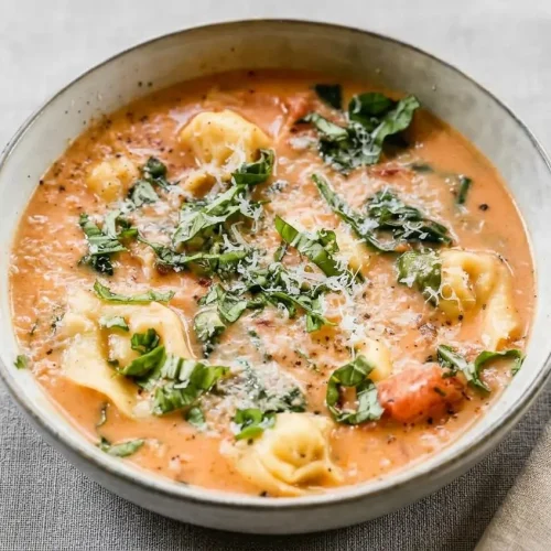 A dark bowl of creamy tomato tortellini soup, garnished with fresh basil leaves and grated Parmesan cheese.