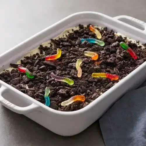 A rectangular pan of Dirt Cake Recipe, showing a crunchy crushed Oreo topping, creamy vanilla pudding filling, and colorful gummy worms.