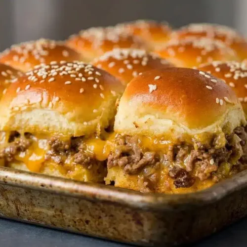 A pan of baked cheeseburger sliders with golden-brown, sesame-seed tops, with one slider being pulled away to show gooey melted cheddar