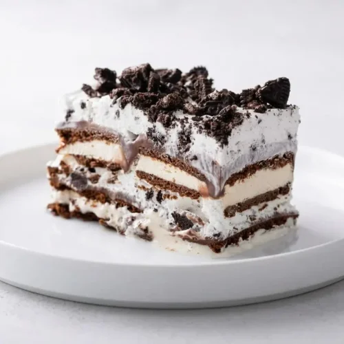 A generous slice of Ice Cream Sandwich Cake on a white plate, showing the distinct creamy layers of fudge, whipped topping, and crushed