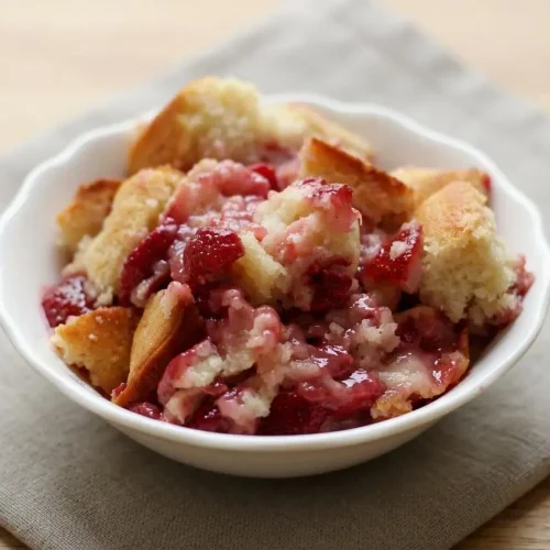 A freshly baked strawberry cobbler in a white oval baking dish, showing its golden brown, fluffy topping and bubbling red berry juices.
