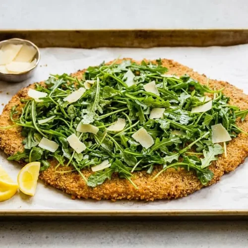 A large, golden-brown Giant Chicken Milanese on a baking sheet, topped with a fresh arugula salad, shaved Parmesan, and lemon wedges.
