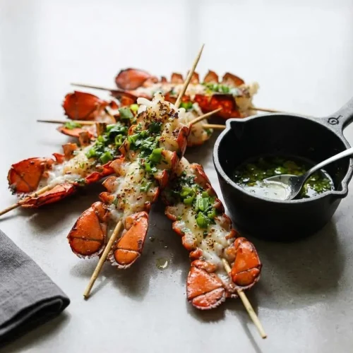 Grilled lobster skewers drizzled with a creamy Bang Bang sauce and topped with fresh cilantro on a dark platter.