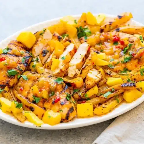 Grilled Mango Pineapple Chicken on a white platter, topped with fresh mango pineapple salsa and cilantro, showing a juicy, smoky finish.