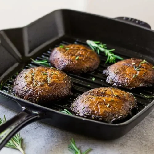Four whole grilled portobello mushrooms arranged on a rustic platter, showing deep grill marks and a glossy balsamic marinade.