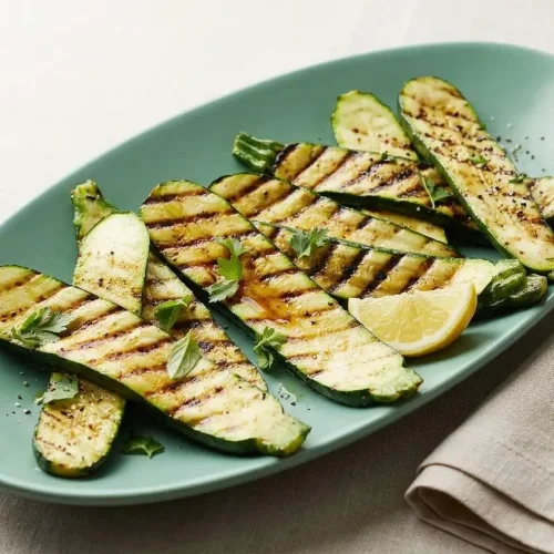 A white rectangular platter of Grilled Zucchini planks with char marks, garnished with fresh parsley and a lemon wedge.