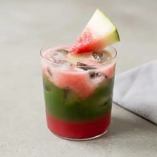 Iced Watermelon Matcha in a tall glass, showing distinct pink and green layers with ice cubes and a watermelon wedge garnish.