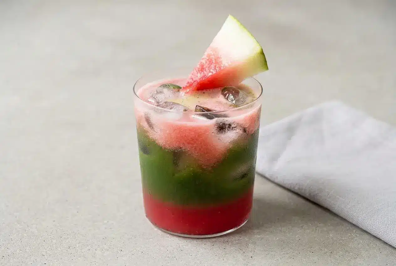 Iced Watermelon Matcha in a tall glass, showing distinct pink and green layers with ice cubes and a watermelon wedge garnish.