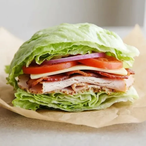 A crisp Lettuce Sandwich wrap cut in half, held in parchment paper, showing layers of turkey, bacon, cheese, tomato, and fresh iceberg
