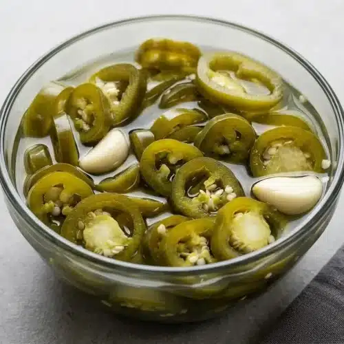 A clear glass jar filled with the finished Pickled Jalapeños recipe, showing sliced green peppers and garlic cloves in a light brine.